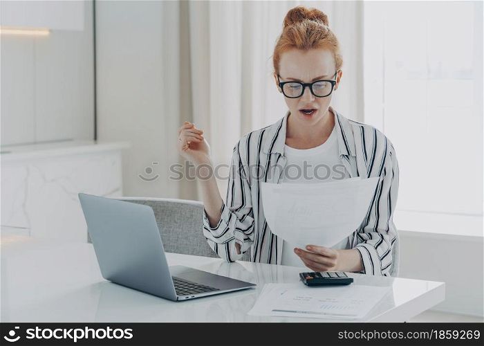 Savings finances concept. Shocked ginger woman focused at document studies bills reads banking paper notification about last morgage payment received letter notice calculates budget sits at desktop. Savings finances concept. Shocked ginger woman focused at document studies bills reads banking paper