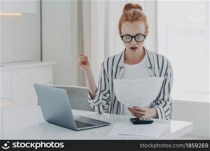 Savings finances concept. Shocked ginger woman focused at document studies bills reads banking paper notification about last morgage payment received letter notice calculates budget sits at desktop. Savings finances concept. Shocked ginger woman focused at document studies bills reads banking paper