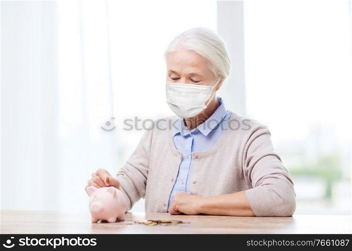 savings, health and old age concept - senior woman wearing face protective medical mask for protection from virus disease putting coin into big piggy bank. old woman in mask putting coin into piggy bank