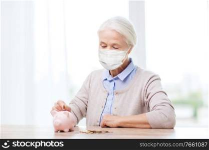 savings, health and old age concept - senior woman wearing face protective medical mask for protection from virus disease putting coin into big piggy bank. old woman in mask putting coin into piggy bank