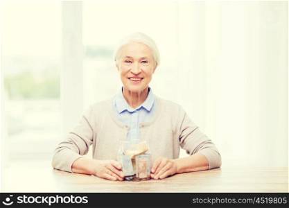 savings, money, annuity insurance, retirement and people concept - smiling senior woman with money in glass jar at home