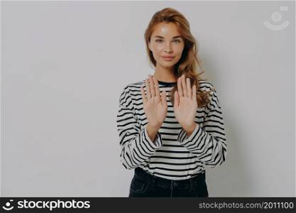Saying no. Young self confident woman holds both hands with palms out and gesturing stop sign, isolated on grey background with copy space for text. Body language and non-verbal communication concept. Young confident woman gesturing stop sign, saying no while standing isolated against grey wall