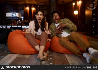 Scared millennial couple feeling fear during watching horror movie on tv projector at home. Scared couple watching horror movie at home