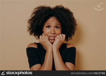 Scared mixed race female with curly hairstyle looking nervously and feeling stress, african woman being afraid of something while posing against beige background. Fear and anxiety concept. Scared mixed race female looking nervously and feeling stress while posing against beige background