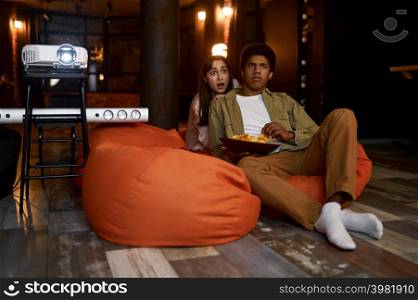 Scared wife hiding behind husband back during watching movie on tv projector together. Young couple watch movie film on projector