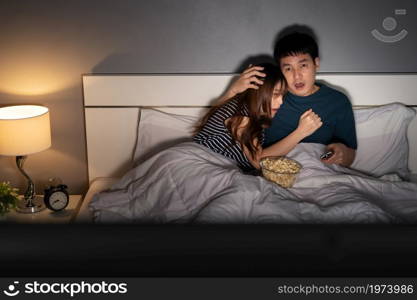 scared young couple is watching horror movie TV on a bed at night