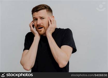 Scared young man in black t-shirt, hands on face. Afraid of horrible scene. Gasping in fright. Isolated on blue studio background. OMG, how scary!