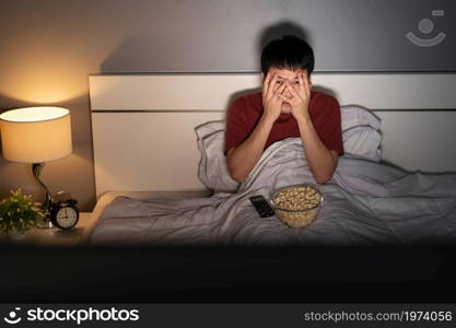 scared young man is watching horror movie TV on a bed at night