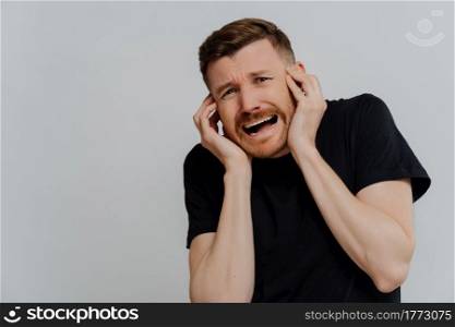 Scared young unshaven man in black tshirt being afraid of something and screaming, looking with frightened expression at something while standing against grey background. Fear and anxiety concept. Scared man screaming while being afraid and looking with frightened expression at something