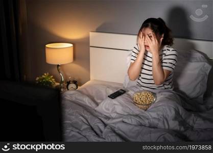 scared young woman is watching horror movie TV on a bed at night