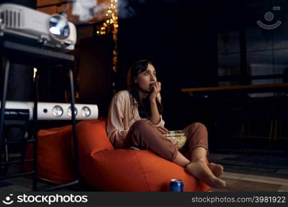 Scared young woman watching horror film using video projector. Leisure activity on quarantine. Scared young woman watching horror film video