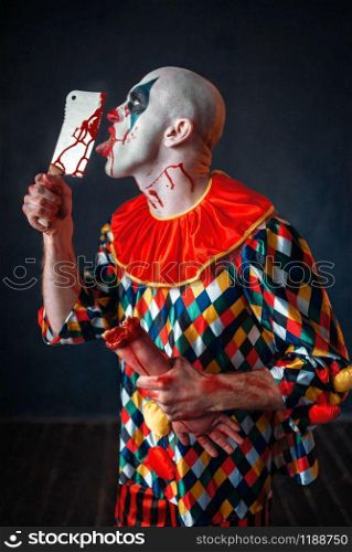 Scary bloody clown licks the knife blade. Man with makeup in halloween costume, crazy maniac holds human hand