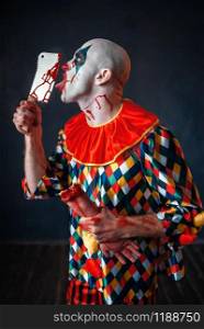 Scary bloody clown licks the knife blade. Man with makeup in halloween costume, crazy maniac holds human hand