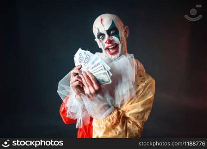 Scary bloody clown with crazy eyes holds fan of money. Man with makeup in carnival costume, mad maniac