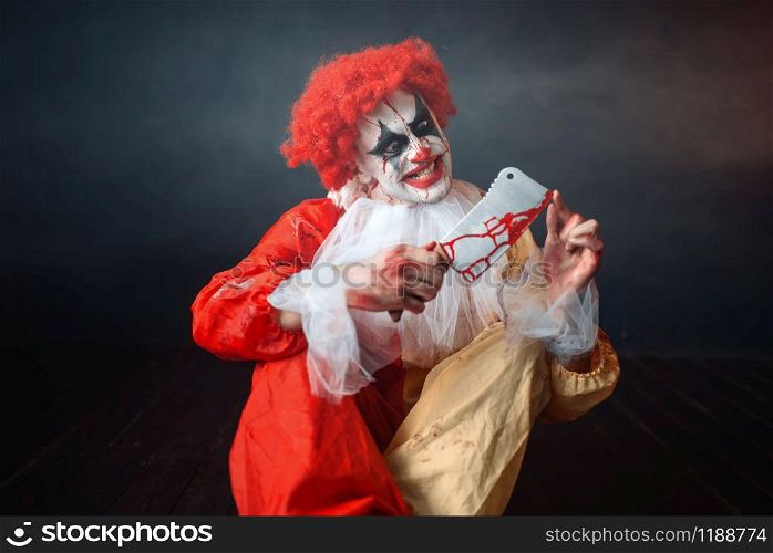 Scary bloody clown with crazy eyes looking on his knife. Man with makeup in carnival costume, mad maniac