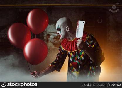 Scary bloody clown with meat cleaver and air balloon sneaking into the basement, horror. Man with makeup in carnival costume, crazy maniac