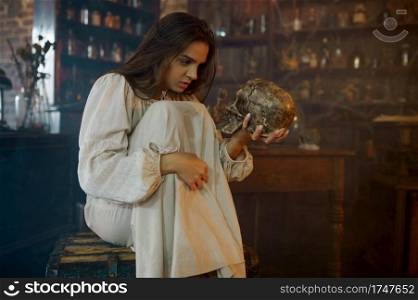 Scary demonic woman holds human skull, demons casting out. Exorcism, mystery paranormal ritual, dark religion, night horror, potions on shelf on background. Scary demonic woman holds human skull, exorcism
