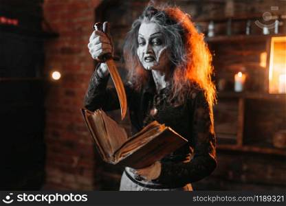 Scary witch holds spellbook and knife, dark powers of witchcraft, spiritual seance. Female foreteller calls the spirits, terrible future teller. Scary witch holds spellbook and knife