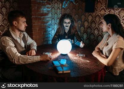 Scary witch reads a magic spell over a crystal ball, young man and woman on spiritual seance. Female foreteller calls the spirits. Witch, man and woman on spiritual seance
