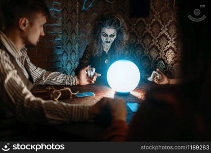 Scary witch reads a magic spell over a crystal ball, young man and woman on spiritual seance.. Witch, man and woman on spiritual seance