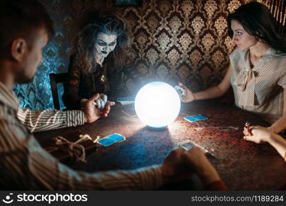 Scary witch reads a magic spell over a crystal ball, young man and woman on spiritual seance. Female foreteller calls the spirits