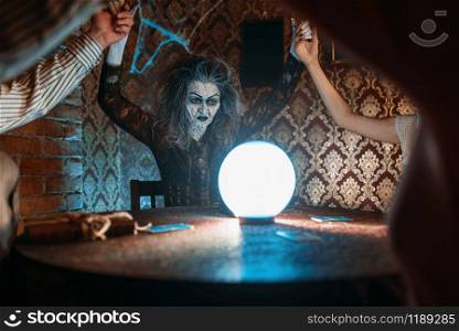 Scary witch reads a magic spell over a crystal ball, young people hands up on spiritual seance. Female foreteller calls the spirits. Witch over a crystal ball, young people on seance