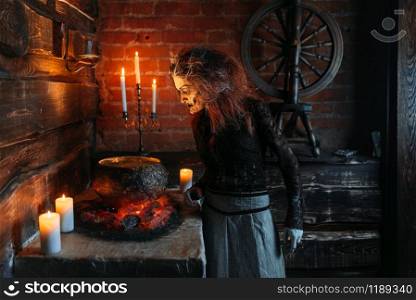 Scary witch reads spell over the pot, dark powers of witchcraft, spiritual seance with candles. Female foreteller calls the spirits, terrible future teller. Scary witch reads spell over the pot, seance