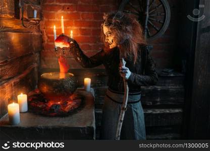 Scary witch reads spell over the pot with human body parts, dark powers of witchcraft, spiritual seance with candles. Female foreteller calls the spirits, terrible future teller