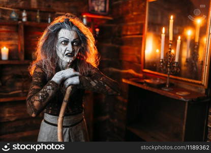 Scary witch with a cane at the mirror and candles, dark powers of witchcraft, spiritual seance. Female foreteller calls the spirits, terrible fortune teller