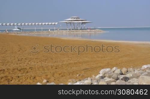 Scene of the resort on Dead Sea. Beach and water view with few people relaxing under the shed in distance