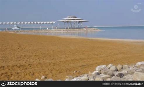 Scene of the resort on Dead Sea. Beach and water view with few people relaxing under the shed in distance