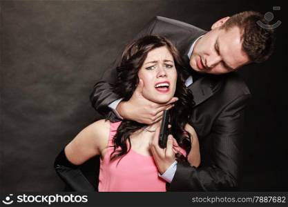 Scene of violence with firearm between men and women. Elegant man holding gun and neck of sitting lady on black and grey background in studio.