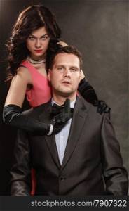 Scene of violence with firearm between men and women. Elegant lady holding gun against sitting man in suit on black grey background in studio.