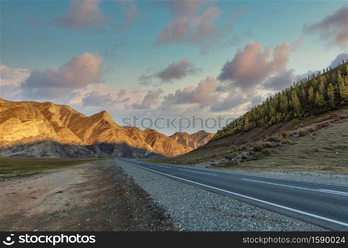 Scenic low angle view of mountain ridge. Highway in the foreground leading all the way up to the mountains. Beautiful cloudy sunset sky as a backdrop. Golden hour. Altai mountains, Siberia, Russia.. Scenic low angle view of mountain ridge. Highway in the foreground leading all the way up to the mountains. Beautiful cloudy sunset sky as a backdrop. Golden hour. Altai mountains, Siberia, Russia