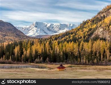 Scenic low angle view of snowy mountain peaks and slopes of North Chuyskiy ridge. Golden trees, wooden hut in the foreground. Beautiful blue cloudy sky as a backdrop. Altai mountains, Siberia, Russia.. Scenic low angle view of snowy mountain peaks and slopes of North Chuyskiy ridge. Golden trees, wooden hut in the foreground. Beautiful blue cloudy sky as a backdrop. Altai mountains, Siberia, Russia