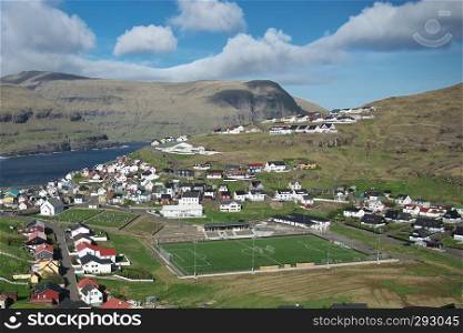 Scenic panoramic landscape of a picturesque village with football stadium and traditional houses on the Faroe Islands. Landscape with mountains, coastline and sky with clouds. Colourful background. Glorious sceneries of the Faroes. Postcard motif.