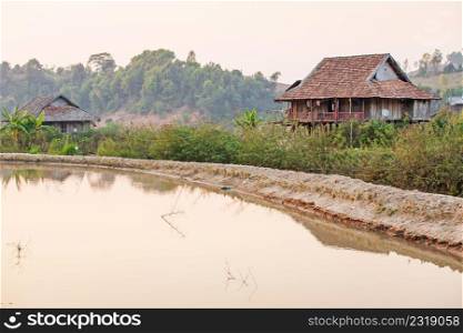 Scenic rural of vietnam at sunset, two old wooden house, brown earthenware roof, pool foreground, forest and mountains backgrounds. Traditional vietnamese architecture house in Dien Bien Phu. Winter season.