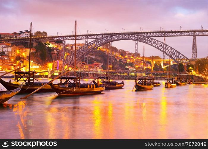 Scenic sunset over Douro river with traditional wine boats, illuminated Porto skyline and Dom Luis I bridge in background, Portugal