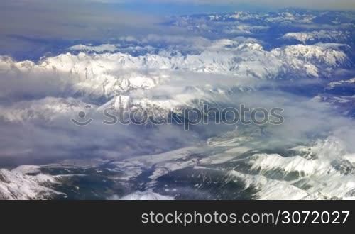 Scenic top view from the flying airplane. Beautiful grand mountains with snowy tops and thin clouds