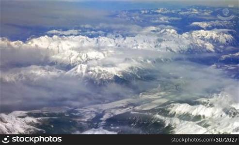 Scenic top view from the flying airplane. Beautiful grand mountains with snowy tops and thin clouds
