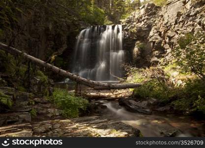 Scenic view of Lower Twin Falls in Glacier National Park with the waterfall set between the rock walls of the stream bed. A long exposure smooths the water and shows the full shape of the falls.