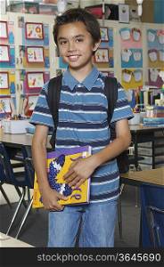 School boy holding book in classroom, portrait
