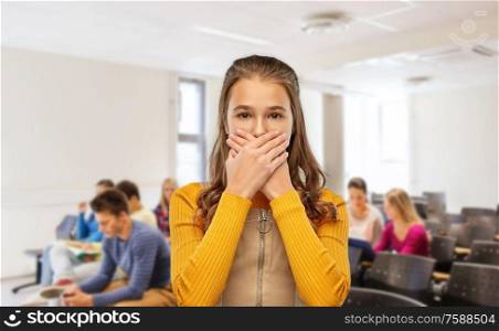 school, bullying and people concept - scared teenage student girl closing her mouth by hands over lecture hall background. scared student girl closing mouth at school