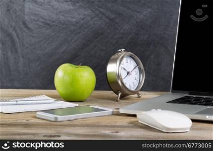 School desktop with laptop, mouse, clock, cell phone, notebook, pen and green apple in front of blackboard. Layout in horizontal format with copy space.