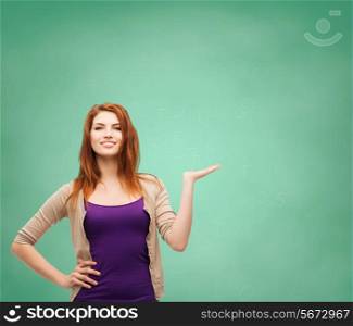 school, education, advertising and people concept - smiling teenage girl in casual clothes holding something on her palm over green board background