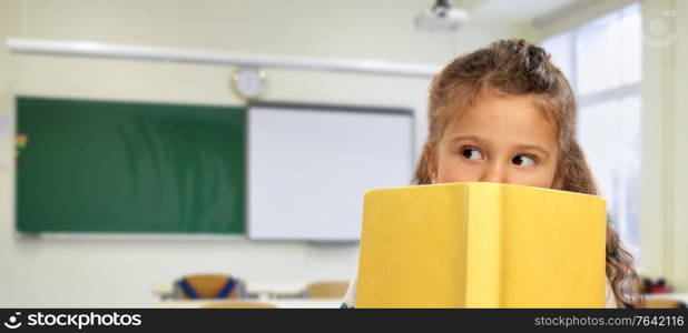 school, education and childhood concept - little girl hiding behind yellow book over classroom on background. little girl hiding behind yellow book at school
