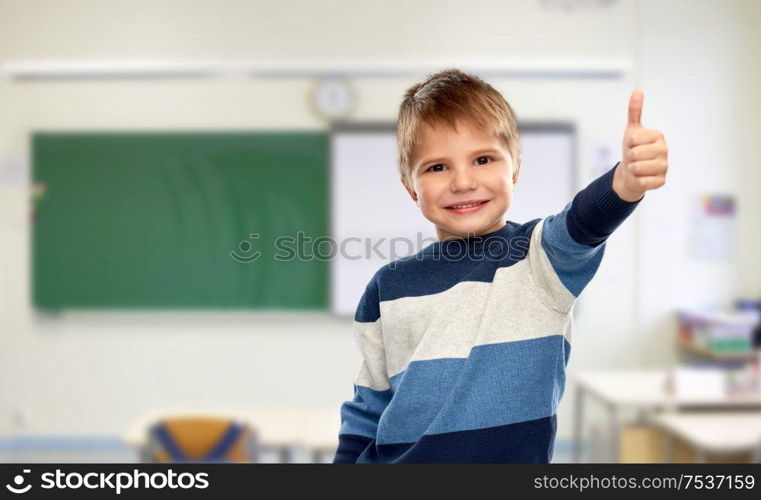 school, education and gesture concept - portrait of smiling little boy in striped pullover showing thumbs up over classroom background. little boy showing thumbs up at school