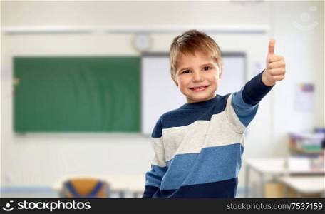 school, education and gesture concept - portrait of smiling little boy in striped pullover showing thumbs up over classroom background. little boy showing thumbs up at school