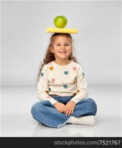school, education and knowledge concept - little girl sitting on floor with book and green apple on top of head over grey background. little girl with book and apple sitting on floor