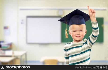school, education and learning and childhood concept - portrait of smiling little boy in mortar board over classroom background. boy in mortar board pointing finger up at school